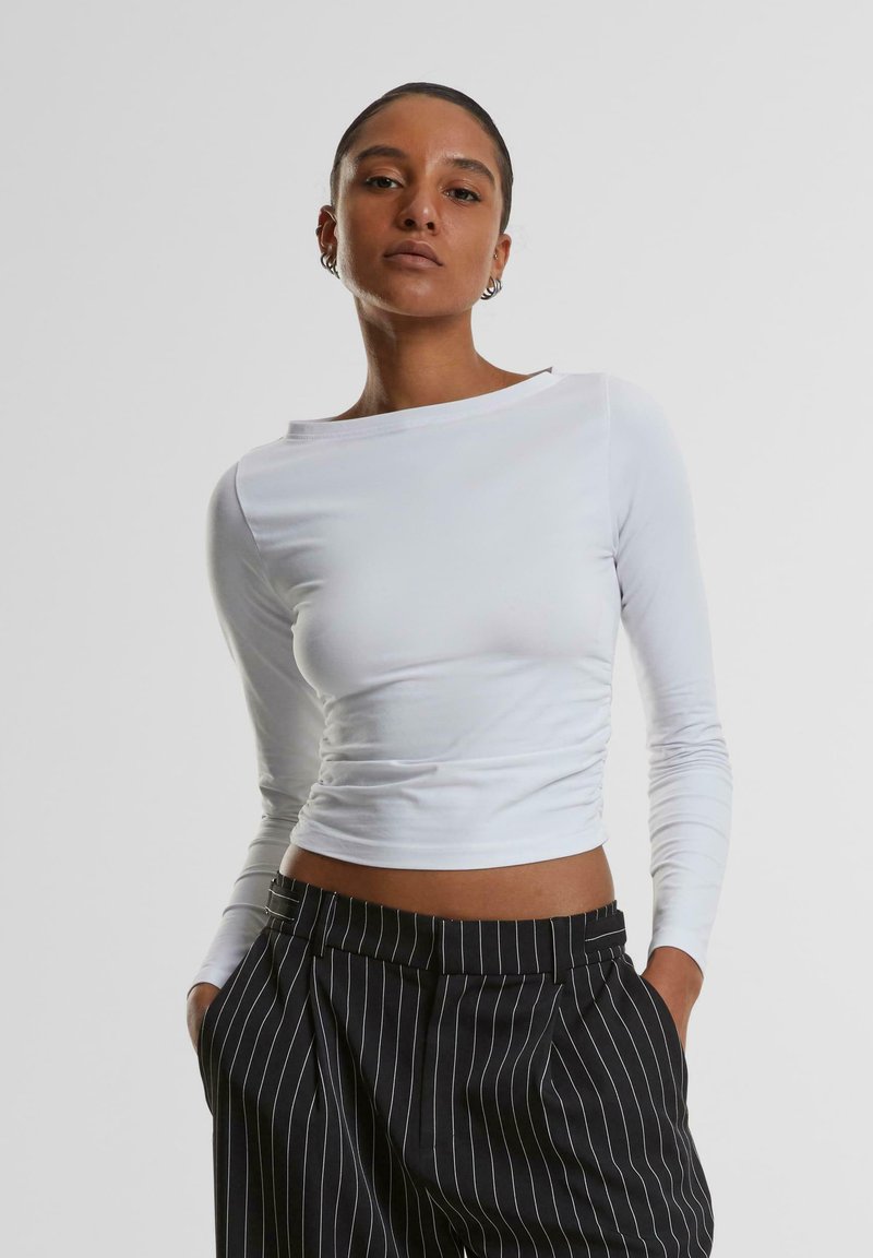 Weißes langärmliges Crop-Top mit Bootsausschnitt und Raffung, kombiniert mit hoch taillierten schwarzen Streifenhosen. Glatte Stoffstruktur.