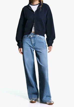 Vrouw die een marineblauwe hoodie met rits draagt, een wit cropped shirt, lichtblauwe wijde jeans en metalen zilveren schoenen, staand tegen een witte achtergrond.