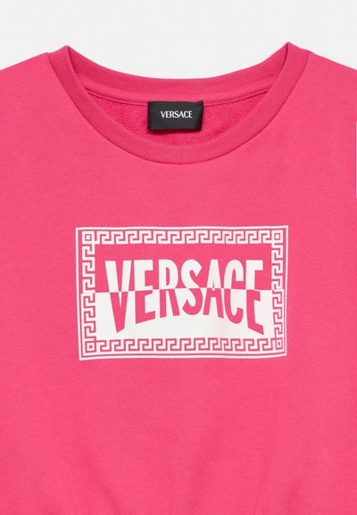 Chemise Versace rose vif avec un logo rectangulaire blanc présentant une bordure géométrique et un texte en gras "VERSACE" au centre.