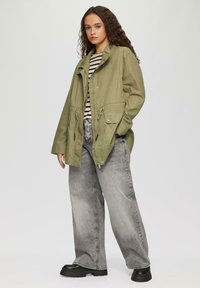 Veste olive avec col montant, fermeture éclair à l'avant et taille ajustable à cordon; associée à un haut rayé et à un jean large gris.