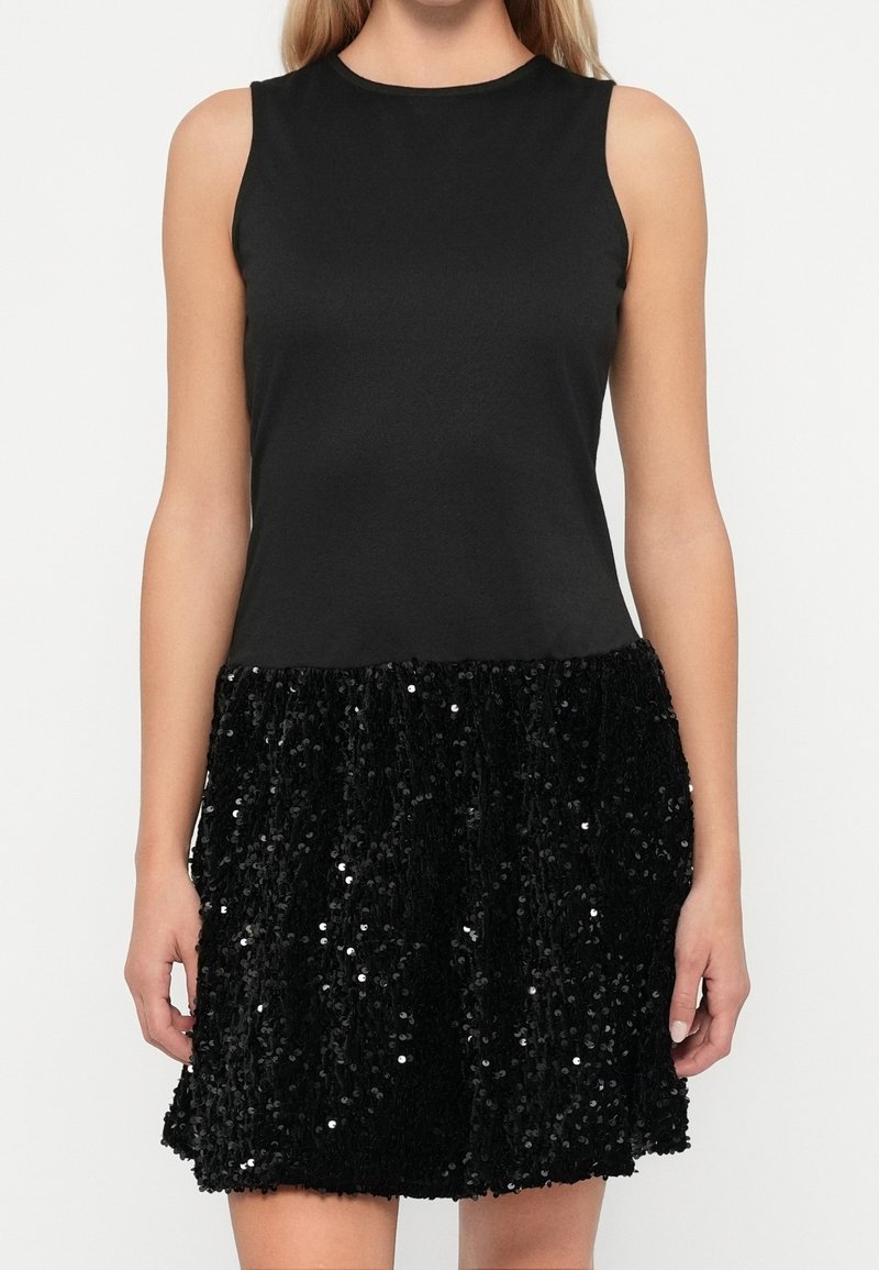 Robe noire sans manches avec un haut ajusté uni et une jupe courte évasée à sequins, portée par une femme debout sur fond blanc.