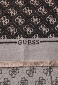 Fekete és krémszínű mintázatú sál, amelyen ismétlődő logódesign található, szilárd krémszínű kerettel és bojt részletekkel. A "GUESS" felirat nyomtatva.