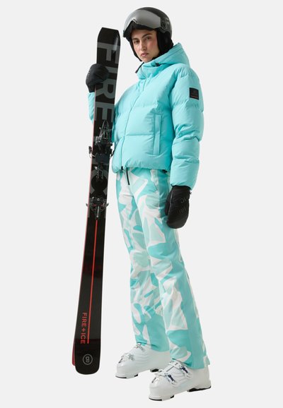 Turkusowa kurtka puchowa z zapięciem na zamek, wzorzyste spodnie narciarskie w jasnoniebieskim i białym kolorze, czarne narty oraz białe buty narciarskie. Czarne rękawice i kask.