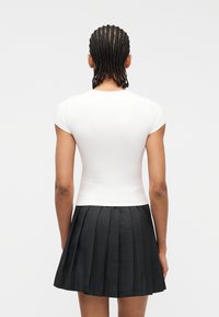 Άτομο με πλεξούδες, φοράει άσπρο εφαρμοστό τοπ με κοντά μανίκια και μαύρη πιετέ μίνι φούστα, εμφανιζόμενο από πίσω.