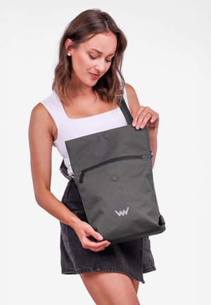 Šedý látkový batoh s horním klopou, zipovou kapsou a logem. Obsahuje nastavitelný popruh a hladkou texturu. Jednoduchý, moderní design.