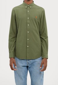 Man i en olivgrön knappad skjorta med långa ärmar och ljusblå jeans, som står mot en enfärgad vit bakgrund.