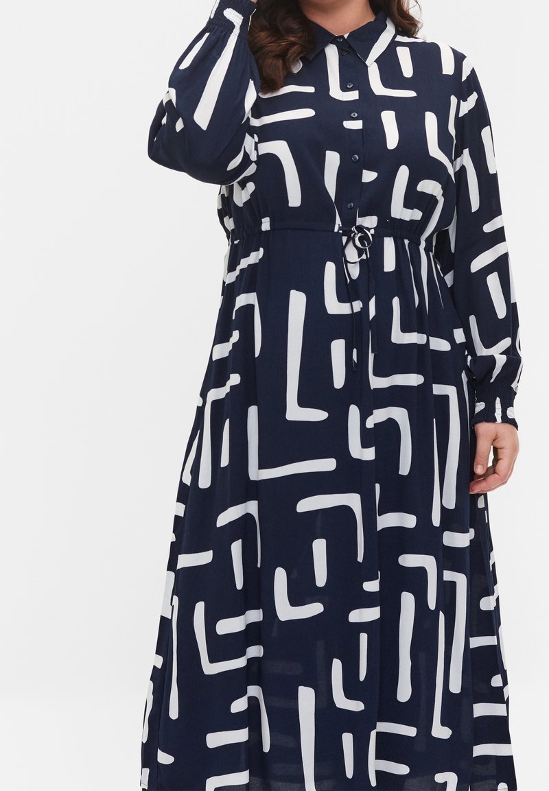 Femme portant une robe midi à manches longues bleu marine avec des lignes géométriques abstraites blanches et une taille à cordon.