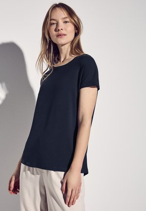 Top nera a maniche corte con scollatura rotonda e vestibilità rilassata, realizzata in un tessuto liscio, abbinata a pantaloni chiari.