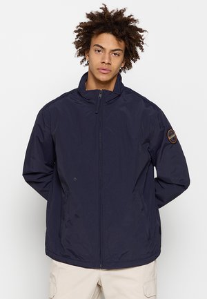 Joven con pelo rizado que lleva una chaqueta azul marino con cremallera y pantalones beige claros, de pie con las manos detrás de la espalda frente a un fondo liso.