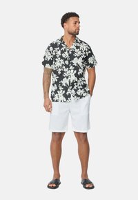 Chemise florale à manches courtes noire avec des fleurs blanches, associée à un short blanc et des sandales noires à enfiler. Offre un design à boutons.