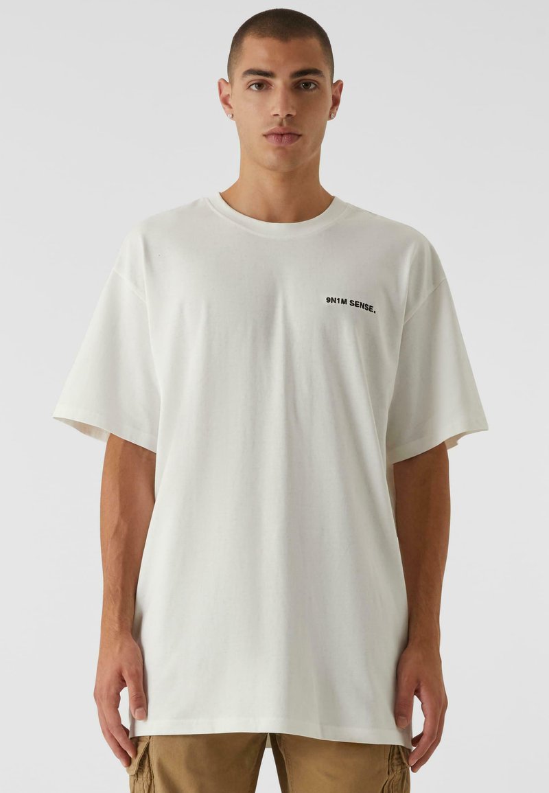 Hvit bomulls T-skjorte med korte ermer, oversized passform. Har en liten svart logo tekst på venstre side av brystet. Glatt tekstur.