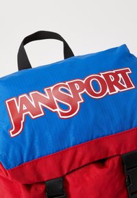 Sac à dos Jansport rouge et bleu avec un grand logo. Équipé d'un tissu lisse et durable, d'un rabat supérieur et de sangles noires.
