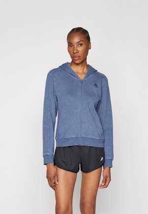 Sudadera azul con cremallera, hecha de una tela suave con un acabado texturizado, que cuenta con una capucha ajustable y tres franjas a lo largo de las mangas, combinada con pantalones cortos negros.