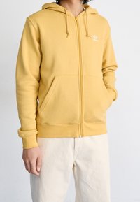 Gul zip-up-huva i mjukt tyg med en framficka, dragsko i huvan och en diskret vit logga på bröstet.