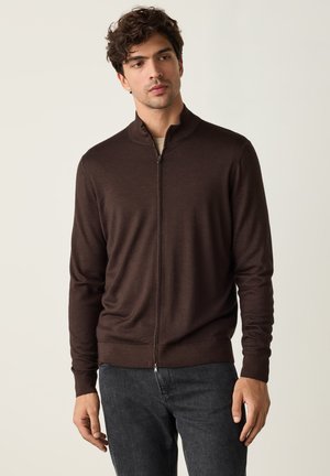 Junger Mann trägt einen dunkelbraunen Reißverschluss-Pullover und dunkelgraue Jeans, steht vor einem einfarbigen hellen Hintergrund.