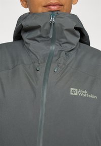 Graue Jacke mit hohem Kragen, Reißverschluss und verstellbarem Kapuzen. Enthält ein Logo auf der Brust und hat eine glatte, wasserabweisende Stoffstruktur.