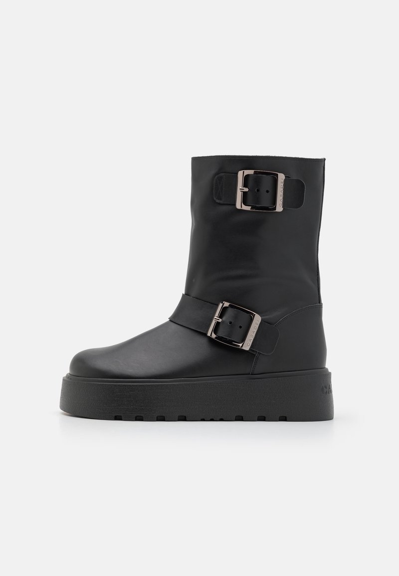 Bottines en cuir noir avec deux boucles en argent surdimensionnées, une semelle épaisse, et une finition lisse et mate.