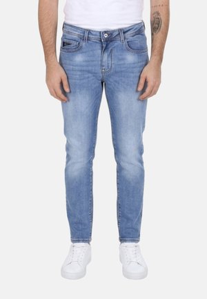 Uomo che indossa jeans skinny azzurri chiari e sneakers bianche, in piedi con le braccia rilassate lungo i fianchi davanti a uno sfondo semplice.