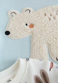 Cream-colored polar bear applique on a light blue background, featuring textured fur, black and orange accents, and white knitted collar.
