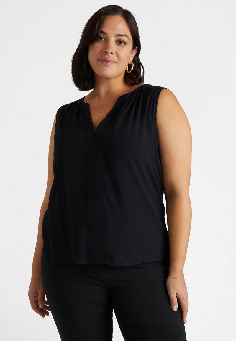 Blusa nera senza maniche con scollatura a V, tessuto morbido, pieghe sottili sulle spalle e vestibilità ampia. Abbinata a pantaloni neri.