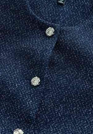 Tengerészkék tweed kardigán texturált szövettel, ezüst csomós knop gombokkal az elején. Nagyított kép a szövet részleteit mutatja.