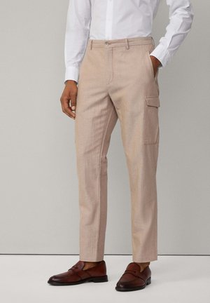 Pantalones cargo de lino beige con un corte ajustado, que cuentan con bolsillos laterales y cierre de cremallera, combinados con zapatos de cuero marrón.