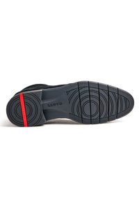 Chaussure avec une semelle en caoutchouc texturée, présentant des motifs circulaires et un accent rouge. La tige est en daim noir avec un design élégant.