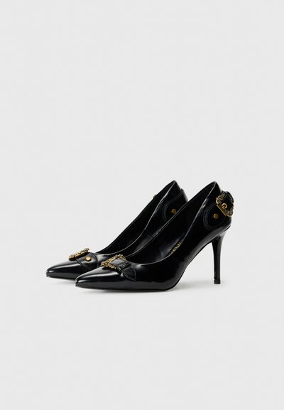 Chaussures à talons hauts en cuir verni noir avec des bouts pointus, présentant un nœud décoratif et des accents en métal doré. Texture lisse.