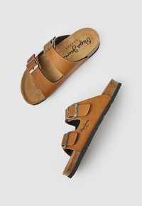 Sandales en cuir marron avec deux boucles métalliques réglables, semelle en liège et semelle en caoutchouc texturée. Logo visible sur la semelle intérieure.