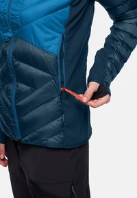 Persona che indossa una giacca blu e navy mentre apre una tasca laterale con una cerniera arancione etichettata "Pack Away".