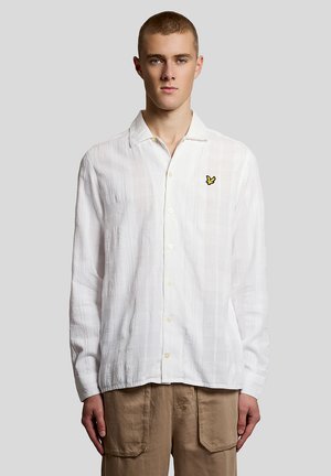 Jeune homme portant une chemise blanche texturée à boutons avec un logo d'oiseau jaune sur la poitrine et un pantalon marron, debout devant un fond uni.