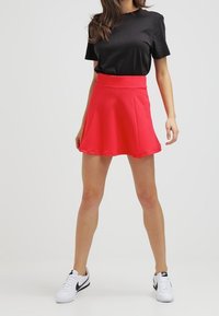 Rode skaterrok van gestructureerde stof, gecombineerd met een zwart t-shirt en witte sneakers. De rok heeft een uitlopende vorm en een middelhoge taille.