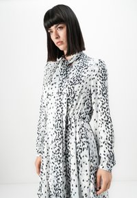 Frau mit glattem, dunklem Haar, die ein langärmliges, weißes Kleid mit schwarzen abstrakten Flecken trägt, stehend vor einem schlichten weißen Hintergrund.