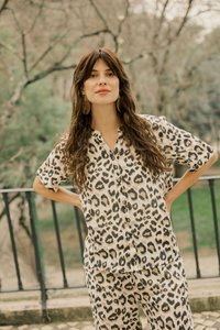 Kvinde med langt brunt hår iført en leopardprint skjorte og bukser står udendørs med hænderne på hofterne nær et sort gelænder.