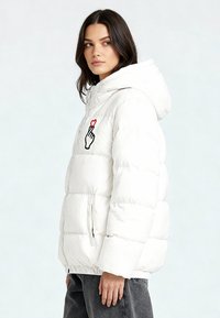 Giacca imbottita bianca con cappuccio, caratterizzata da un ricamo di una mano nera e un cuore rosso sul petto e tasche laterali per riporre oggetti.