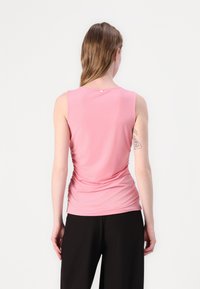 Rosa ärmlös topp med ruchdetaljer vid sidorna och en liten accentknapp på baksidan. Slät tyg, figurnära design, bärs med svarta byxor.