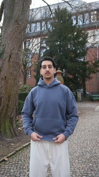 Joven de pie sobre un camino empedrado, viste una sudadera azul con capucha y pantalones blancos, con un gran árbol y un edificio de ladrillo al fondo.