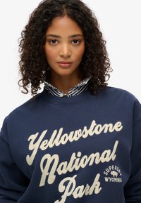 Sudadera de marino con la inscripción "Parque Nacional Yellowstone" en letra crema, con un cuello a rayas visible debajo. Fabricada con tela suave.
