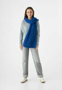 Jeune femme debout, souriante, portant un pull gris, un jean gris clair, des baskets blanches et une longue écharpe en laine bleue.