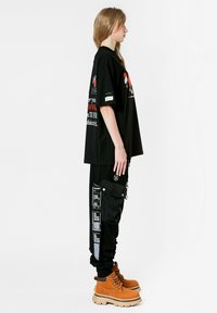 Camiseta negra con estampado gráfico, combinada con pantalones cargo negros que tienen parches y un bolsillo adjunto. Botas marrones con suelas gruesas.