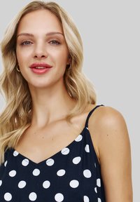Blondynka z falującymi włosami, nosząca złote kolczyki w kształcie kół oraz granatowy bezrękawnik w duże białe kropki, delikatnie się uśmiechająca.