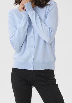 Personne portant un cardigan bleu clair boutonné et un pantalon noir, les mains jointes près du visage sur un fond uni.