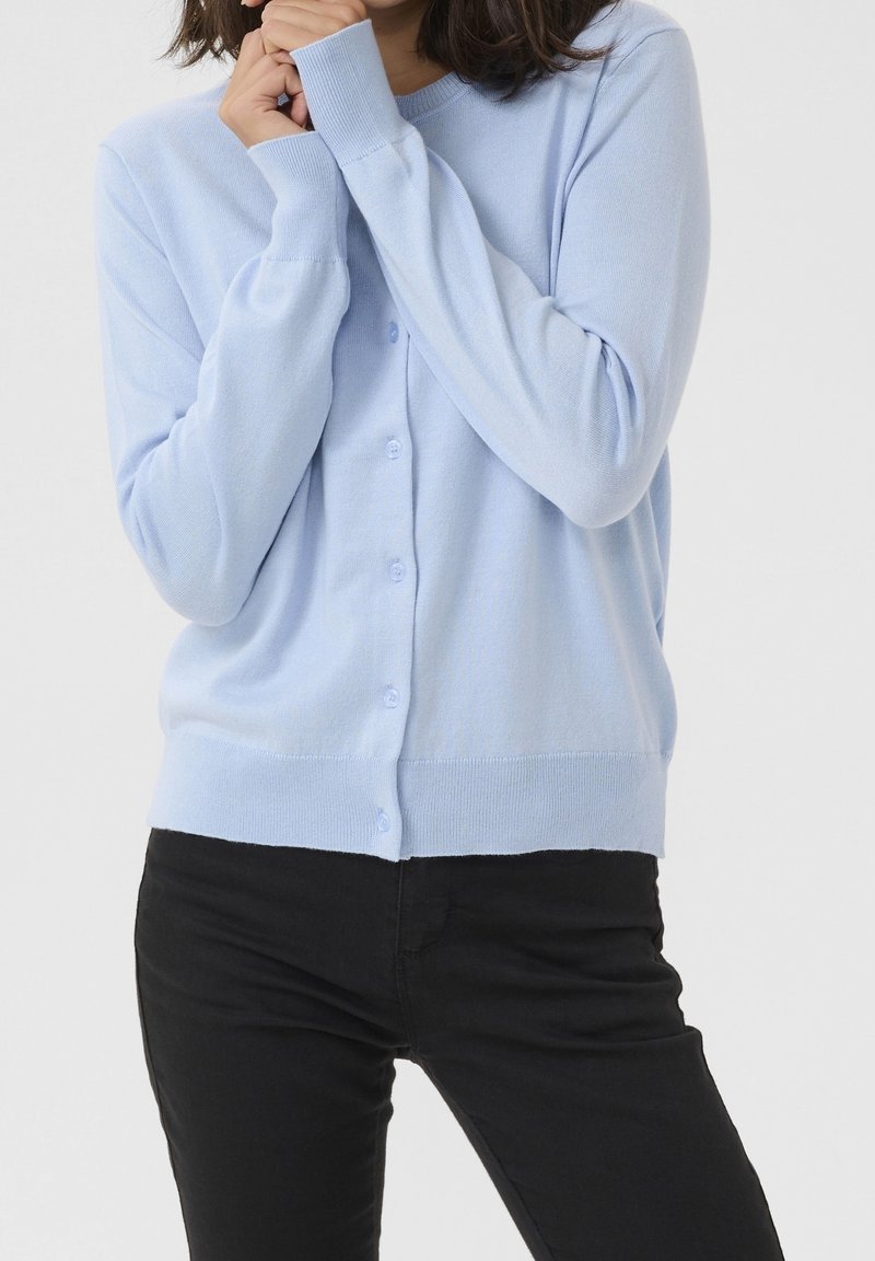 Personne portant un cardigan bleu clair boutonné et un pantalon noir, les mains jointes près du visage sur un fond uni.