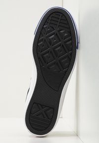 Semelle de chaussure en caoutchouc noir avec un motif de crampons multidirectionnels, des parois latérales blanches et des accents texturés. Présente le logo sur la semelle extérieure.