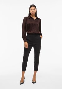 Smeđa saten košulja s dugim rukavima i gumbima, u kombinaciji s crnim klasičnim hlačama i crnim štiklama s zlatnim akcentima.