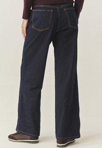 Tmavě modré džíny se širokými nohavicemi a žlutým prošitím, dvěma zadními kapsami a hladkou texturou látky, doplněné hnědými teniskami.