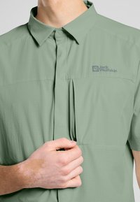 Korte mouwen, lichtgroen shirt van een textuurstof met een geknoopt kraag, voorzien van een ritszak en een logo op de borst.