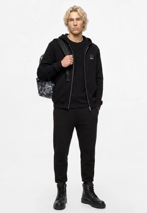 Jeune homme aux cheveux blonds portant un sweat à capuche noir, un pantalon et des bottes, portant un sac à dos marbré noir et blanc sur une épaule.