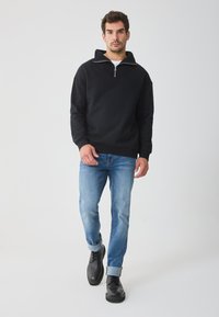 Sweatshirt preto com fecho éclair e capuz, apresentando textura canelada na gola e nos punhos. Combinado com jeans azul claros e botas pretas.