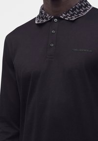 Polo noir à manches longues avec trois boutons noirs et col à motif, avec logo embossé "KARL LAGERFELD" sur la poitrine.